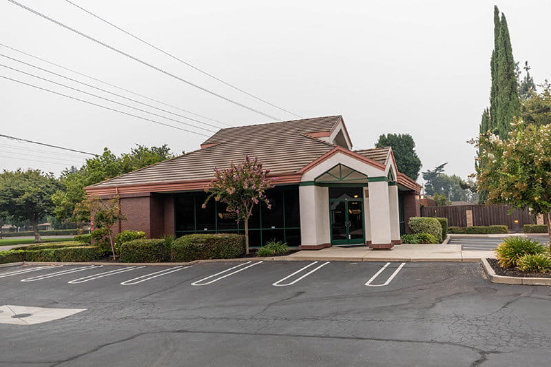 Building from parking lot. White building with red roof, lots of bushes.